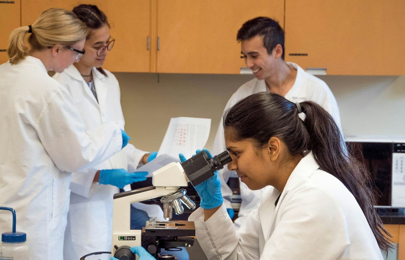 Four laboratory professionals in white coats working together around a microscope
