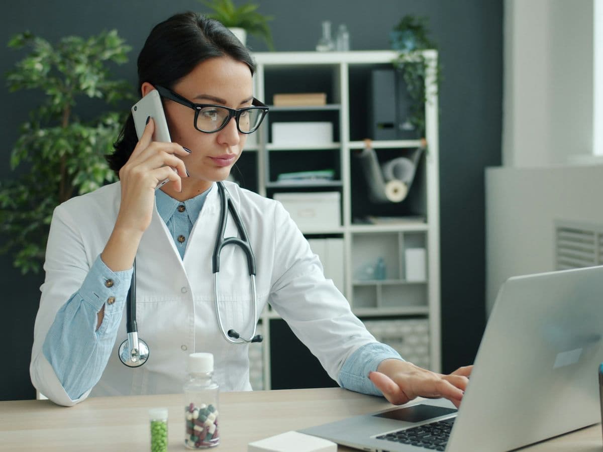 Doctor in a white coat speaking on a phone while working at a laptop