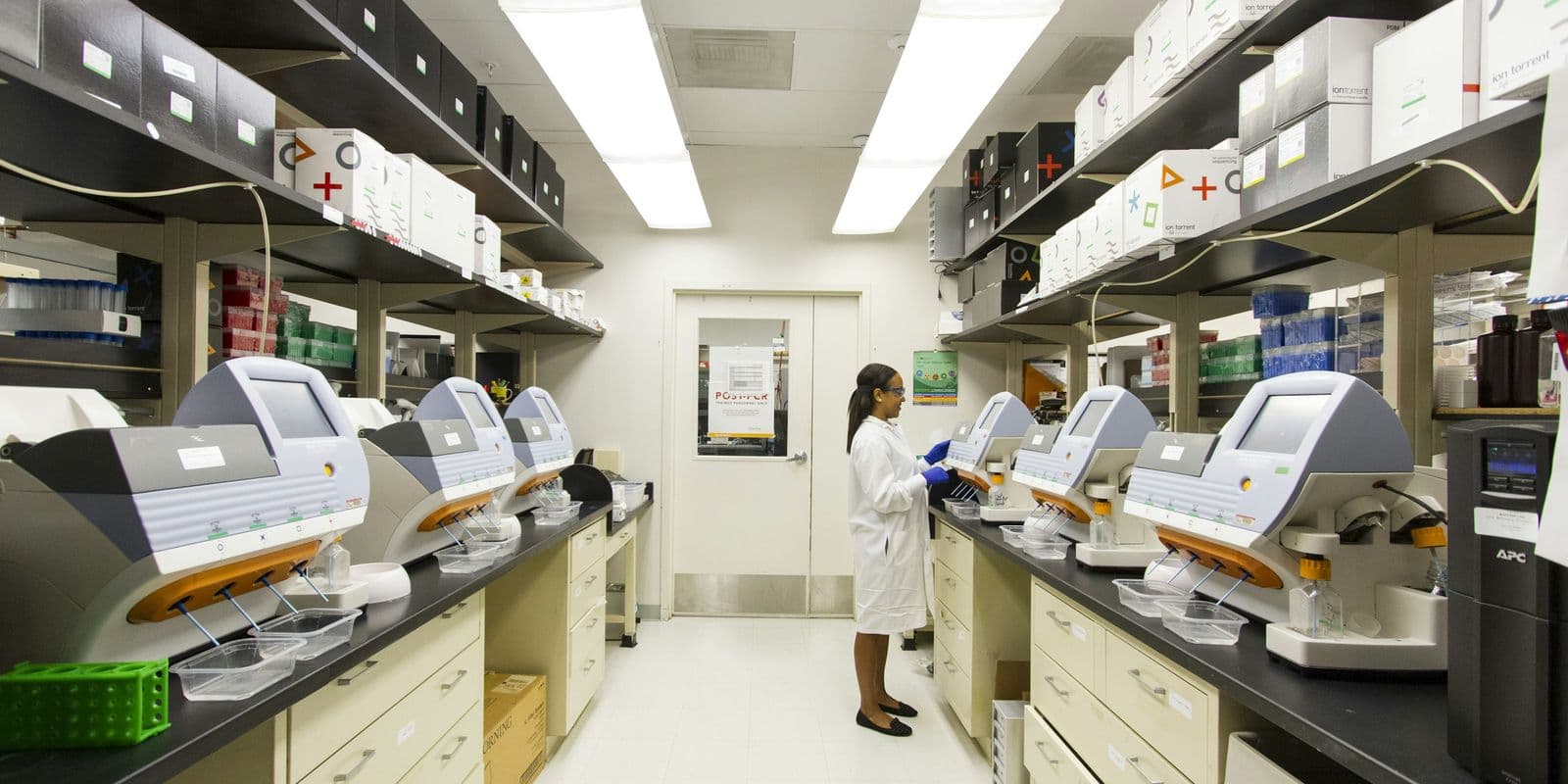 Long bright laboratory aisle with analyzers on benches and a technician in a white coat